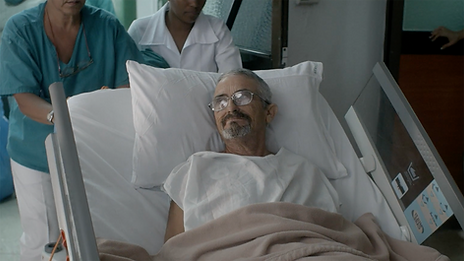 An elderly trans man lying in a hospital bed. He is wearing wire-rimmed glasses and a white hospital gown, and has short, white-streaked grey hair, and grey facial hair. Two nurses are wheeling his bed through a hospital hallway.
