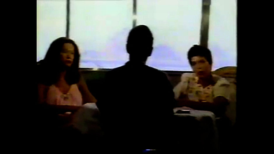 Solange Bastos, a Brazilian journalist, interviewing João W. Nery on TV Manchete. João is sitting in the foreground, a black silhouette, with none of his features visible. He is facing away from the viewer. Sitting in the middle ground are Solange (a brown-skinned, slender woman with short, dark hair) and Joana (a brown-skinned, slender woman with long, dark hair). The background is blurred windows comprised of bright green and vivid blue colours, due to the footage being old.