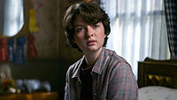 Theo Putnam, a trans boy in Chilling Adventures of Sabrina, played by Lachlan Watson, wearing a flannel shirt and sitting in his bedroom. He has short, wavy black hair and light skin.