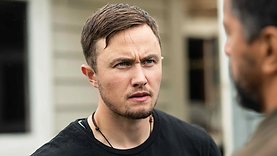 Caz, a trans man played by Māori trans male actor Elz Carrad, in the trans male movie Rūrangi. He is standing outdoors, looking at something with a determined or stern expression. He is lighter-skinned with short, brown hair, and brown stubble. He is wearing a black shirt and a leather necklace. A house is visible in the blurred background.