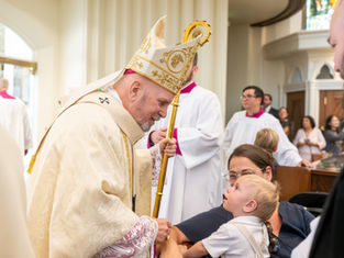 Bishop in gold vestments and mitre smiles and greets a young child held by his mother during a church liturgy, with clergy and parishioners in the background.