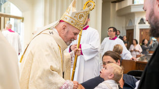 Bishop in gold vestments and mitre smiles and greets a young child held by his mother during a church liturgy, with clergy and parishioners in the background.