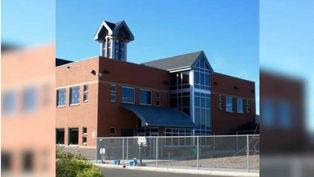 Red-brick building with large glass windows and tower against blue sky. Fenced yard in front, suggesting a secured area or campus.