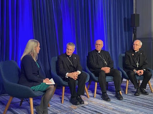 A woman interviews three men in clerical attire seated on blue chairs, against a blue curtain backdrop. The setting is formal and calm.