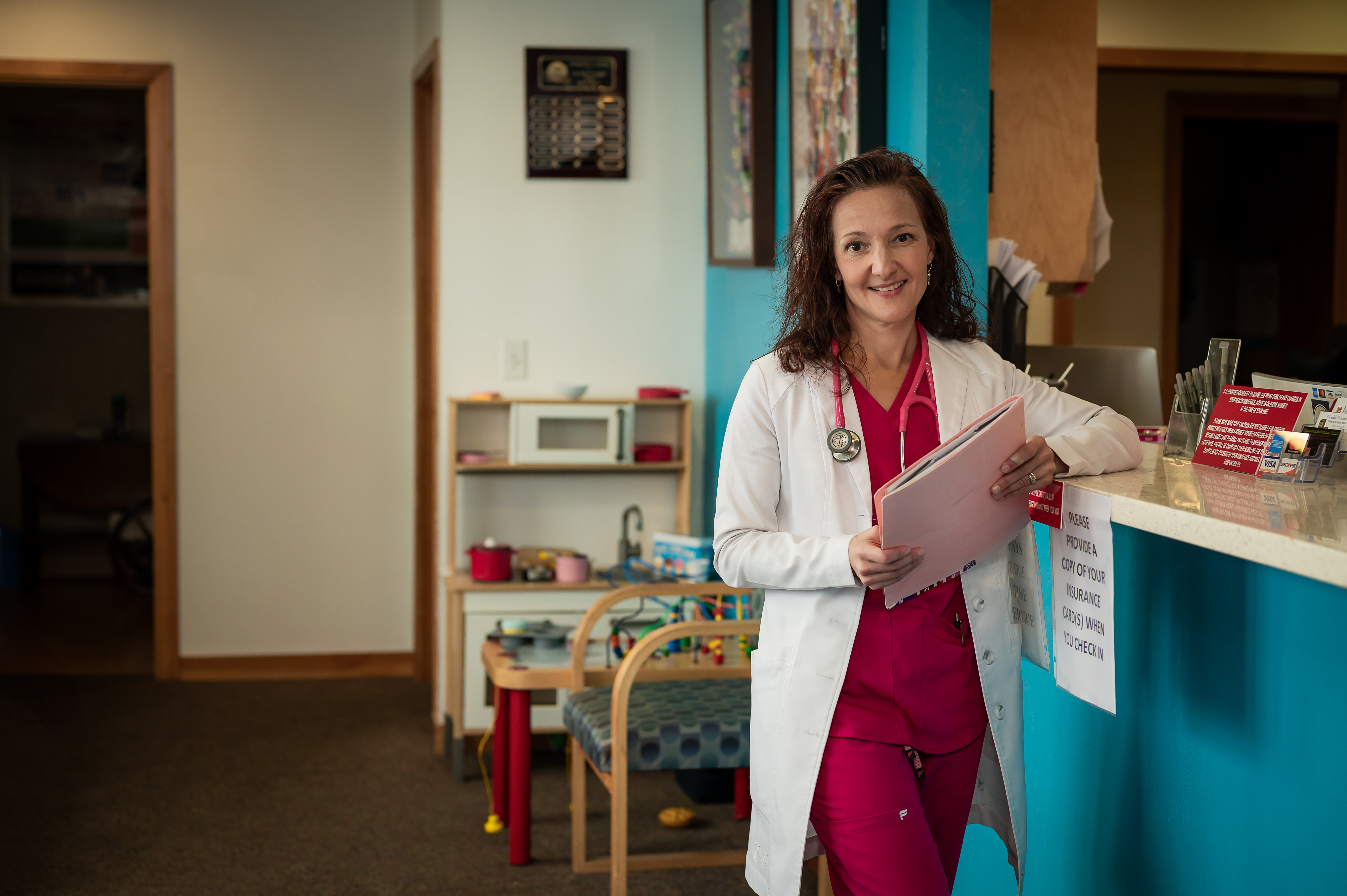 Female doctor smiles, wearing lab coat, holding a chart, leaning near counter.