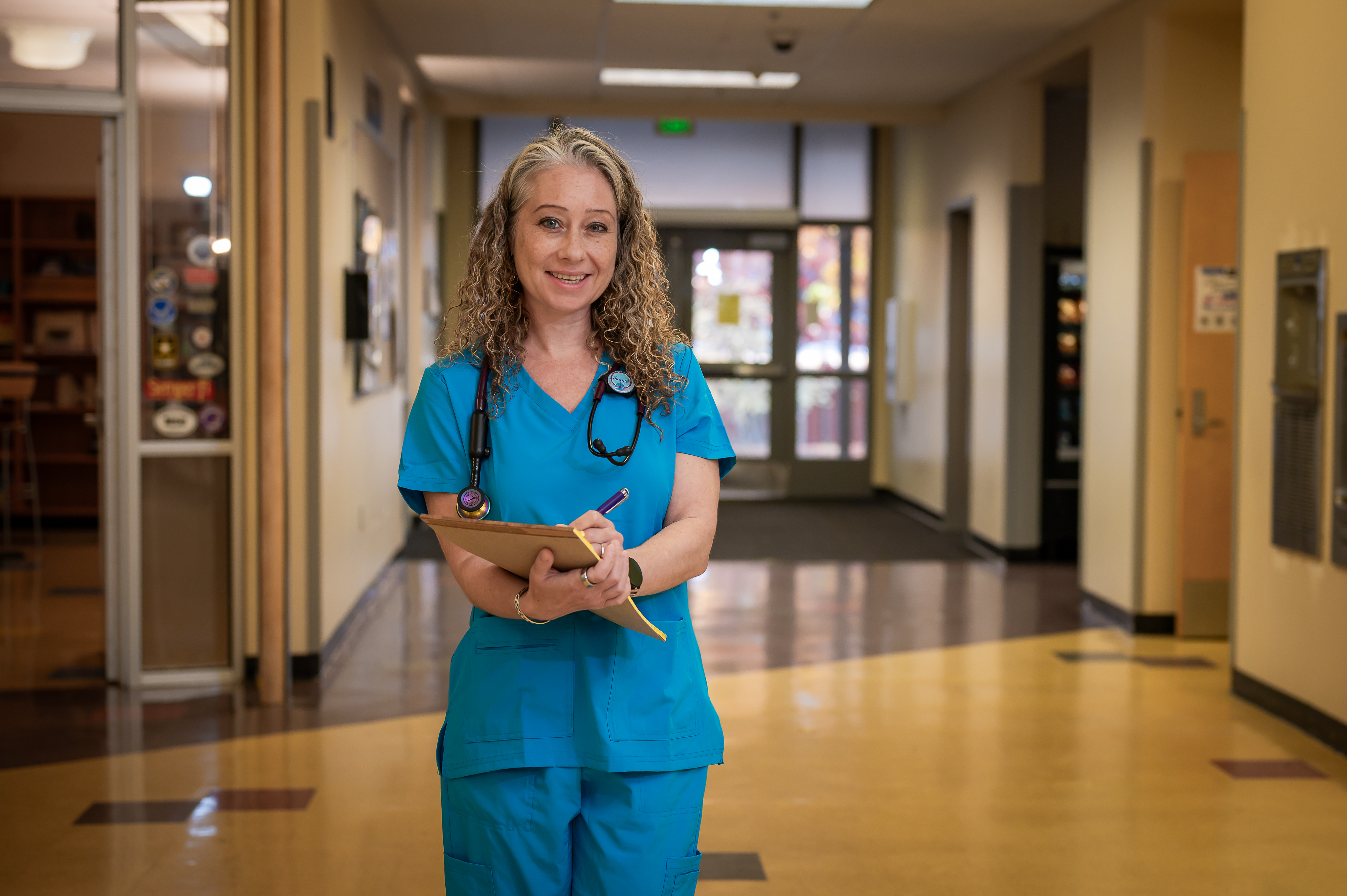 Smiling nurse holding clipboard stands in a well-lit hospital hallway.