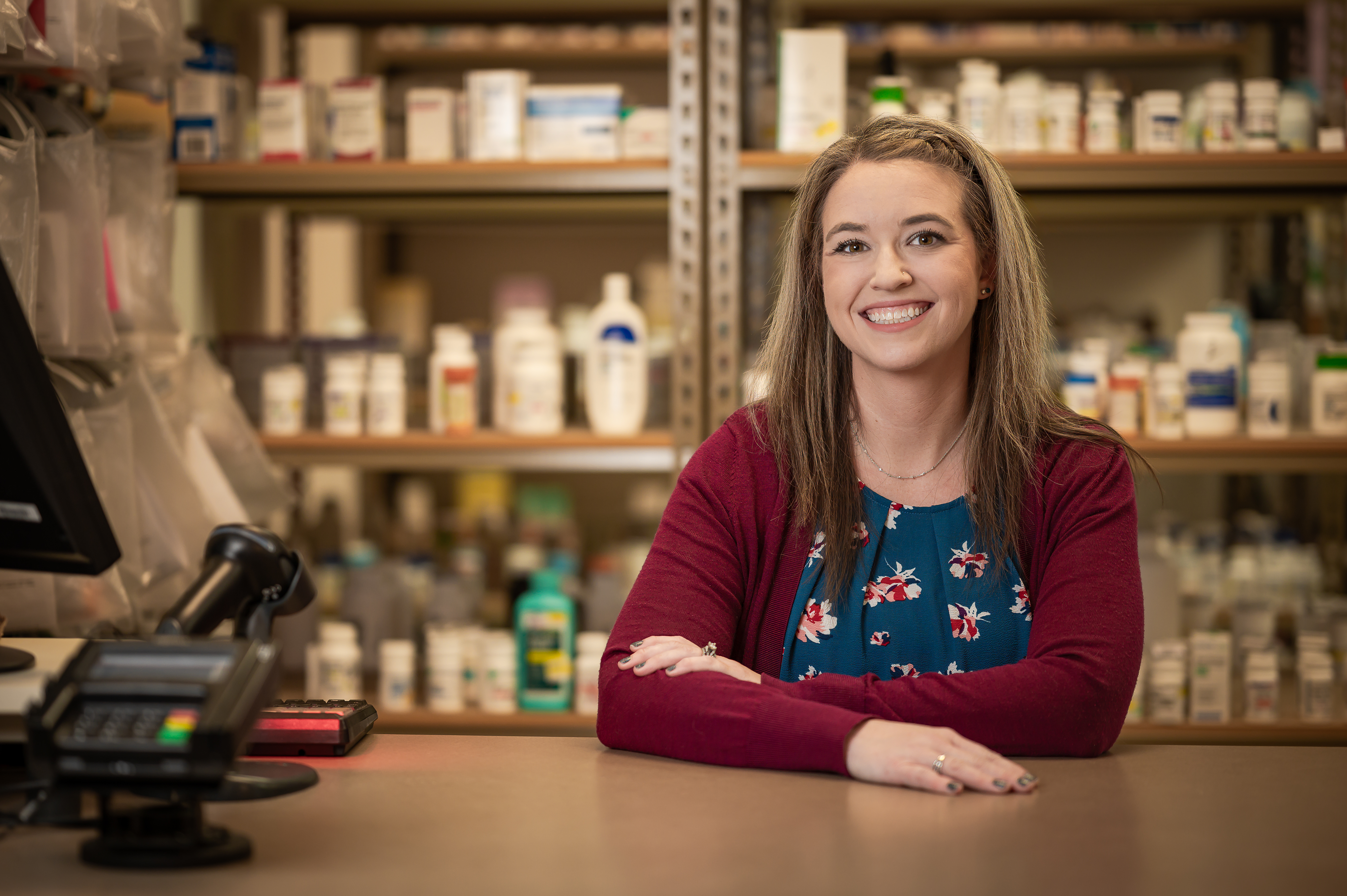 Smiling pharmacist standing in a pharmacy, many medications on shelves behind.