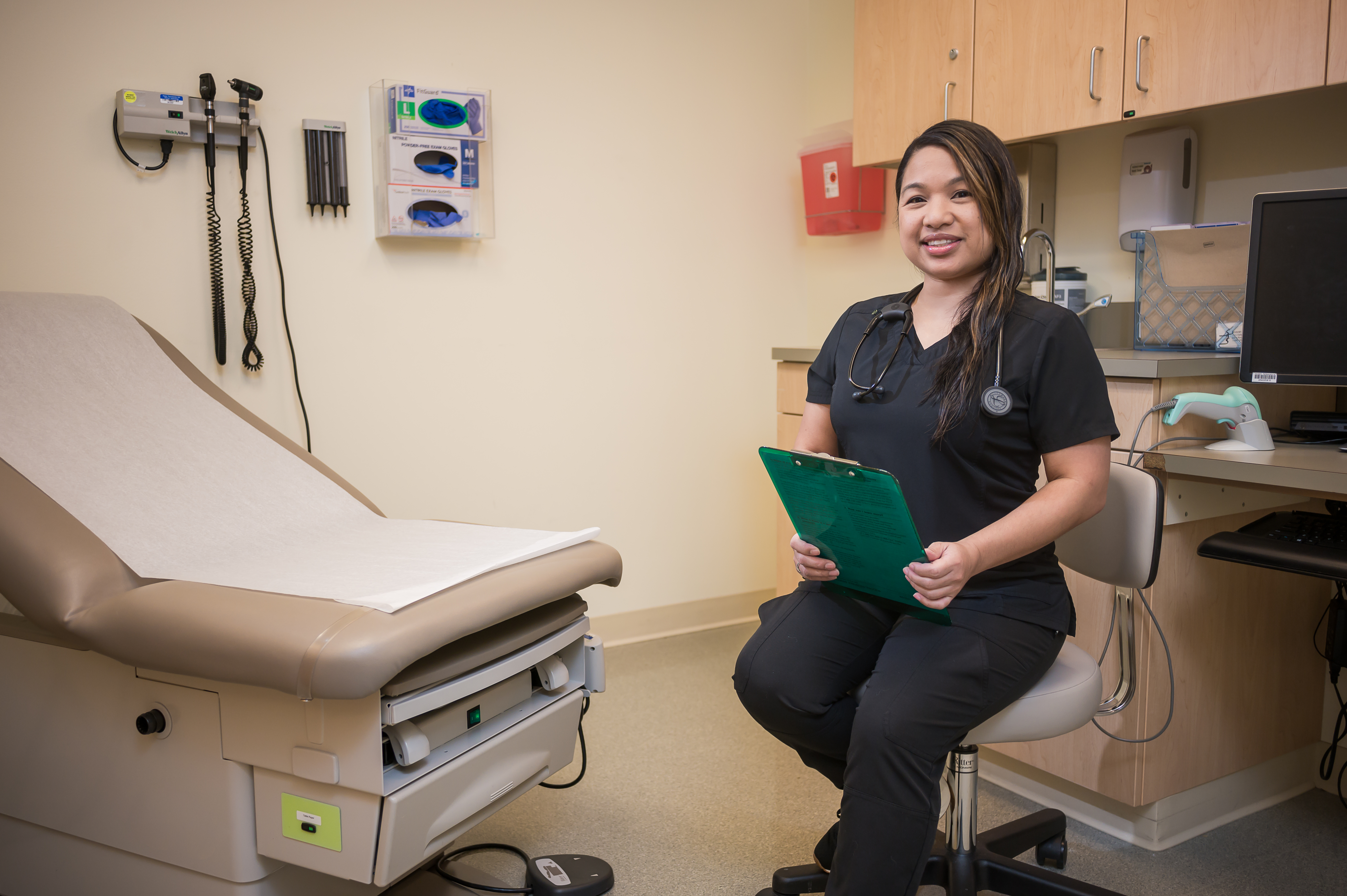 Smiling medical professional holding a clipboard in an examination room setting.