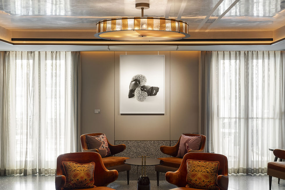 Elegant lounge with rust velvet chairs around a small table, abstract art, sheer curtains, and a gold-lit ceiling chandelier.