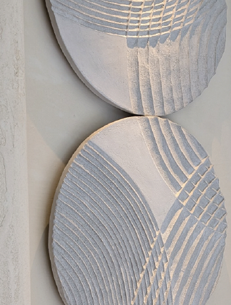 A close-up shot shows a pair of textured, round wall art pieces with ridged patterns, presented against a neutral, off-white wall. The lower piece occupies the primary visual space and exhibits a design featuring a white background overlaid with a motif comprised of textured ridges in a light gray hue. These ridges form concentric arcs near the bottom, transitioning to straight lines at the top right.  The top piece is positioned partially out of frame with only the bottom visible and mirrors the texture and coloration of the lower segment. The ridges on both pieces are not uniformly colored, showing subtle variations in the light gray, which adds depth and dimension.  The texture is visibly rough, which accentuates the three-dimensional quality of the artwork. Light reflects off the ridges and the flatter surfaces, highlighting the texture and the interplay of light and shadow in the design. The close proximity of the shot reveals the intricacies of the material and the construction of the pieces, underscoring the handcrafted or artisanal appearance of the decor.