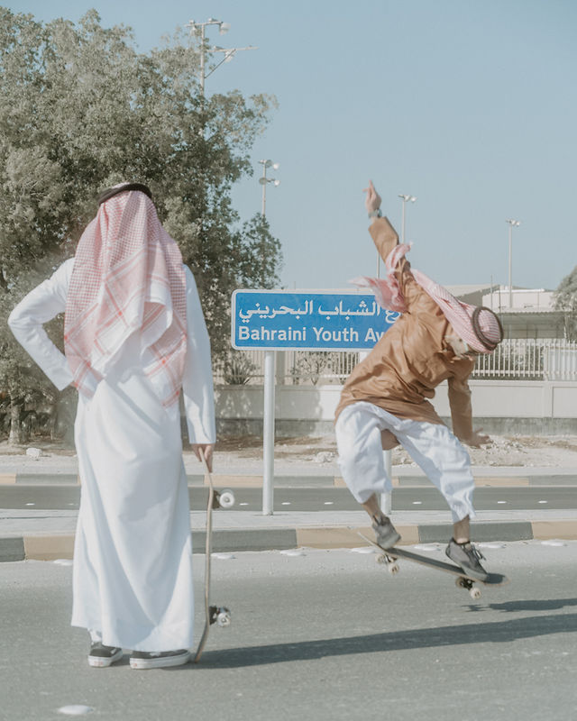 Two men in traditional attire skateboarding on a sunny street near a "Bahraini Youth Avenue" sign, conveying a blend of tradition and modernity. Photograph by Ishaq Madan