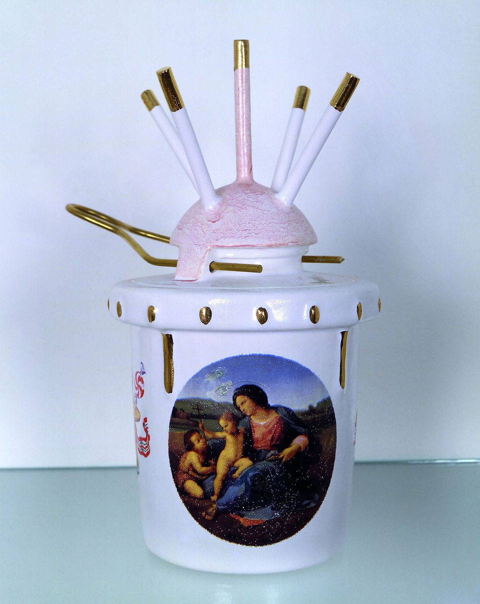 A decorative, cylindrical ceramic object is centered in the frame, displaying a combination of artistic and functional elements. The object is light colored, primarily white, and adorned with golden accents. It features a print of a classical painting or artistic rendering, presumably depicting religious or historical figures, enclosed within an oval frame.  The ceramic structure is topped with a pink, umbrella-like attachment featuring several protruding rods and a central cylindrical component extending upwards. These rods are white with gilded tips.  The base of the object is decorated with repetitive patterns of short, vertical lines, also in gold. Golden tools or accessories are attached or inserted into the sides of the ceramic piece.  The backdrop is a plain, light-colored wall that provides a neutral setting, ensuring the ceramic object remains the focal point. The lighting is diffused, casting soft shadows and evenly illuminating the piece, which aids in showcasing the details and contours of the artwork and the object's overall structure.