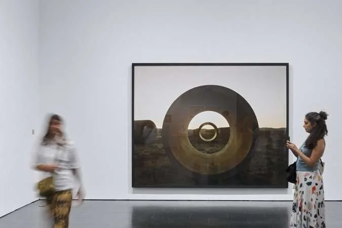 In a brightly lit, indoor setting, an eye-level shot captures a large framed picture prominently displayed on a white wall. The picture, encased in a thin black frame, features a surreal landscape. There's a juxtaposition of circles within circles, creating a tunnel-like effect. The outer circle is large and imposing, with a smaller circle visible through it, and yet another, even smaller circle nested within that. The landscape visible through the circles is barren and somewhat desolate, with a muted color palette of browns, grays, and a hint of pale yellow.  Slightly blurred in the foreground, on the left side of the frame, is a person walking by. Their attire consists of a light-colored top and patterned pants in shades of yellow and brown. The blur suggests movement, indicating they are in transit through the gallery or exhibition space.  The floor beneath is sleek and reflective, mirroring the room and the artwork to a subtle extent. This adds depth and dimension to the composition. The overall mood is contemplative, with the surreal nature of the artwork inviting introspection.