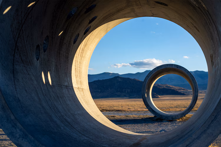 The image captures two concrete industrial pipes standing in the middle of a vast, somewhat barren field. The pipes are arranged in such a way that the larger one frames the scene, with the smaller pipe visible through its opening, set slightly to the right and further in the distance. The interior of the larger pipe is bathed in warm sunlight, which creates patterns of light and shadow on the surface. These patterns are further enhanced by the presence of several evenly spaced, oval-shaped holes in the upper section of the pipe's interior.  Beyond the pipes, the landscape stretches out towards a range of dark, rugged mountains silhouetted against a light blue sky. The sky is partly clear, with a few scattered wisps of clouds adding texture and depth. The field itself consists of dry, tan vegetation, and the ground immediately around the pipes appears to be a rough, concrete-like surface. The overall composition is a striking blend of industrial and natural elements, creating a sense of scale and perspective.