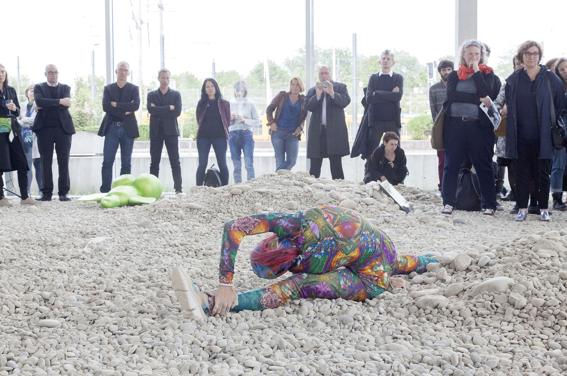 In a medium shot, a person in a vibrantly patterned, full-body suit performs calisthenics on a pile of white pebbles in what appears to be an art installation or performance piece. The individual, distinguished by their pink hair, is positioned with one leg stretched forward and the other to the side, their torso bent forward, and their hand resting near their foot.  The attire is a riot of colors—blues, greens, purples, oranges, and pinks—arranged in a dense, floral-like pattern that fits snugly against the person's body. The suit's fabric appears slightly synthetic and has a subtle sheen, suggesting a material designed for flexibility and comfort. The person's shoes are light-colored and athletic, providing a practical contrast to the artistic display.  A blurry mass of lime green material is visible to the left of the individual, likely another installation element. Behind the pebble mound, a group of casually dressed spectators stand and observe. They’re a diverse mix of ages and styles, with some wearing jeans and t-shirts, while others are in more formal attire like jackets and button-down shirts. One person is actively taking a photo with a mobile device, indicating engagement with the scene.  The background reveals the interior of a modern building, evidenced by the large windows and minimalist design. Natural light filters through, illuminating the space and highlighting the texture of the pebbles. The overall atmosphere suggests a temporary or unconventional setting for this public display.
