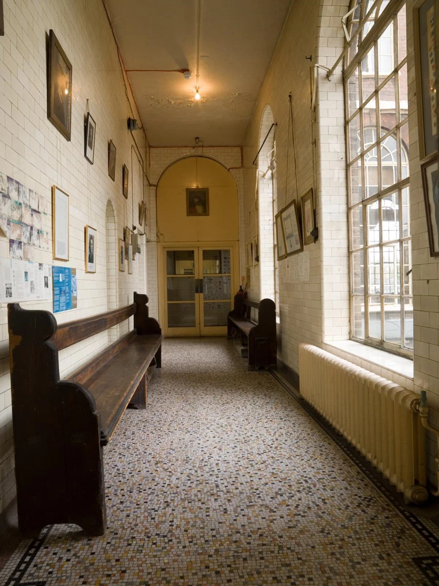 The picture shows a long, slightly aged hallway with old-fashioned decor. On the left, a wooden bench with a high, sculpted back rests against a wall covered in white, rectangular tiles. Various framed pictures and posters are affixed to the wall above the bench.  Across the hall, on the right, are tall windows divided into numerous panes, allowing natural light to illuminate the space. A vintage radiator sits beneath the windows, and above it, more framed pictures adorn the wall.  The floor is composed of small, multi-colored tiles, and the perspective of the hallway leads to a pair of closed double doors at the far end. Above the doors, the wall is painted with a muted yellow hue and features an arched design, with a framed portrait hanging centrally. The electrical wiring is visible along the ceiling, which is somewhat worn and aged. A simple light fixture hangs from the ceiling, casting a warm glow over the hallway.