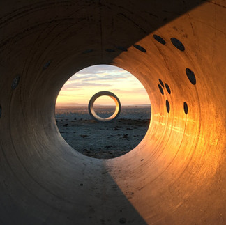 The image shows a view through a large concrete or metal pipe at a desert landscape with a circular sculpture in the distance. The pipe's interior is textured and unevenly lit, with the right side catching the warm light of the setting sun, while the left is cooler. A series of dark, oval cutouts or marks are visible on the right side of the pipe's interior.  Through the opening of the pipe, the landscape of the desert extends and reveals another, smaller circular sculpture standing upright on the horizon line. The sky is a soft gradation of color, indicating either sunrise or sunset, with light clouds adding depth and texture. The overall mood of the scene is tranquil and serene, with the framing of the pipe creating a sense of perspective and focus in the vast, open space.
