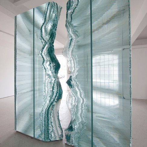 A slightly low-angle, medium shot features an impressive, vertical glass structure split in the middle and standing on a neutral-toned floor in a bright, white-walled room. The structure is comprised of numerous thin, horizontal panes of glass, stacked neatly on top of one another, creating a block-like design.  The split in the middle of the structure is notably organic, with edges that appear as if they have been sculpted or eroded. These organic edges create a visual contrast to the geometric regularity of the horizontal panes. The glass structure appears to act as a partition or decorative wall, adding an artistic element to the modern, minimalist room.  The room is brightly lit, and the light permeates through the glass structure, illuminating the fine details of the texture and layers. Through the transparent gap in the split structure, one can see another part of the room, with a window featuring a grid pattern, further adding to the geometry of the scene. The overall ambiance suggests a modern art installation presented in a clean, uncluttered setting.