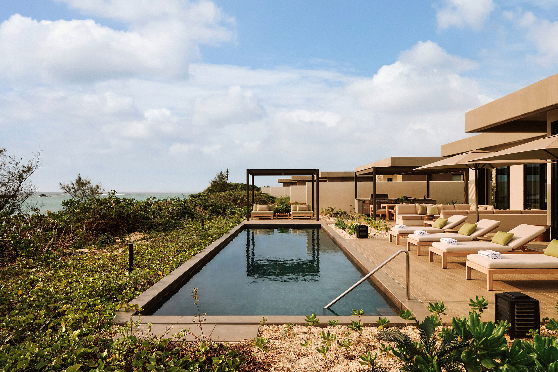 The eye-level, wide outdoor shot displays a serene vacation setting with a rectangular in-ground pool in the foreground, flanked by vegetation and opening to a view of the ocean in the distance.  The pool's dark water surface reflects the soft blue sky, dotted with fluffy white clouds. A simple, modern pergola with square posts and a flat roof stands beyond the pool, sheltering two lounge chairs with soft green cushions.  To the right, a wooden deck is furnished with an array of lounge chairs, also adorned with green cushions and neatly folded white towels. Behind the lounge chairs, outdoor sofas with similar green cushions are positioned under the shade of large umbrellas. A modern outdoor kitchen is visible in the back, complete with a grill and dining area, all part of a contemporary, sand-colored building that merges with the natural environment.  The pool is bordered by a light-colored stone and surrounded by lush, green vegetation, with a small section of sandy beach visible on the left, leading towards the ocean. The view of the ocean is partially obscured by thick shrubs and trees. The sky above features scattered clouds that create a soft, diffused light.