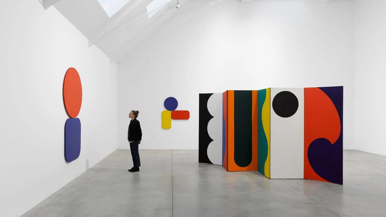 A woman stands in a modern art gallery, observing a colorful array of artworks on display. The gallery space is characterized by its whitewashed walls, high ceilings, and a polished concrete floor. White fabric hangs from the ceiling.  To the right, a series of tall, rectangular panels stand hinged together, each painted with different geometric shapes and vibrant colors. The first panel is black with three white semicircles stacked vertically. The adjacent panel is white. The next panel features a bold orange outline around a dark green center, with a touch of purple, and the following panel showcases a wavy yellow stripe against a vibrant teal background. The final panel in this series is white with a large black circle at eye level.  To the left, on the white wall, a smaller composition of geometric shapes is mounted. It includes a blue circle placed above a yellow rectangle with rounded corners, and a red, rounded rectangle extending from the right side of the yellow shape.  The woman, dressed in dark jeans, a black jacket, and black shoes, is positioned on the left side of the frame, facing the artworks. Her posture suggests contemplation as she regards the art, emphasizing the gallery's focus on the viewer's interaction with the displayed pieces. A small bun in her blond hair sits at the back of her head.