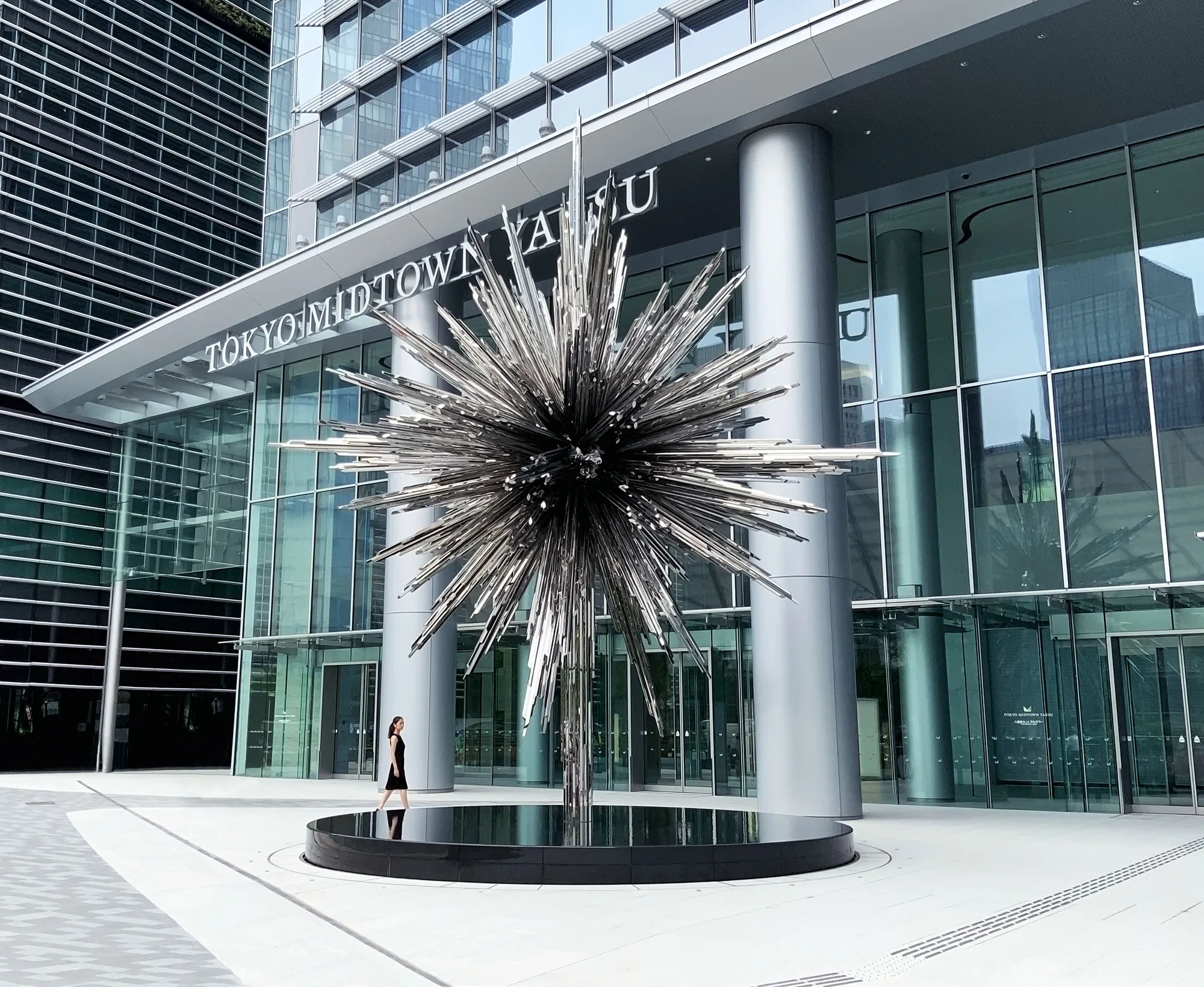 58. © Photo by CASACOR showing a large metal sculpture installation outside a corporate space called Tokyo Midtown in grey and blue tones with a woman in a black dress walking left to the artwork which looks like a metal star or asteroid