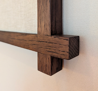 A closer-up, eye-level shot showcases the intersection of two dark-brown-colored wooden boards. The boards are rectangular, with one running horizontally and overlapping a vertical board. Each board's exposed ends show square corners. Light reflects off the rich wood grain, highlighting the texture. The boards frame a neutral, off-white canvas, or cloth material in the artwork. The background is plain, light-colored wall.