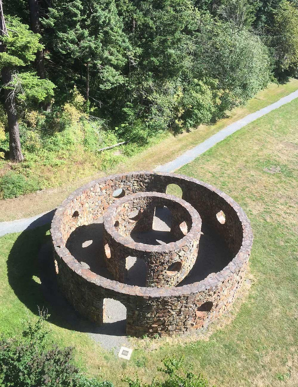 A high-angle shot captures a unique stone structure nestled in a park-like setting. This artwork consists of two concentric circular walls made of rough-cut stones. The smaller, inner ring is enclosed by the larger, outer ring, creating a spiral-like path between them. Both rings have several uniform, circular gaps in their walls, giving the structure an open and airy feel.  The ground inside and around the stone structure is covered in well-manicured grass. To the upper right of the frame, a well-maintained path can be seen, winding its way through the green landscape. Beyond the path, a dense forest of lush green trees forms a natural backdrop.  The lighting suggests a bright, sunny day, casting soft shadows that accentuate the texture of the stone and the contours of the landscape. The overall composition creates a sense of harmony between the man-made structure and the natural environment.