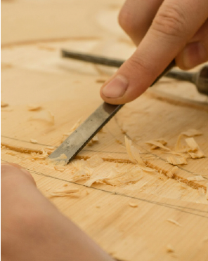This up-close image shows the careful act of wood carving. At the heart of the scene is a woodworker's hand, gently holding a steel chisel. The blade of the chisel is pressed against a light-colored wooden surface, meticulously carved along a drawn line. Tiny wood shavings curl and scatter around the carved area, a testament to the focused work in progress.  The surface of the wood around the carving is marked with faint pencil lines, guiding the artisan's hand. In the background, a handle-less wood chisel subtly comes into view, mirroring the tool being used. The shallow depth of field keeps the focus firmly on the hand, the chisel, and the carved area, emphasizing the detailed and delicate nature of the woodworking.