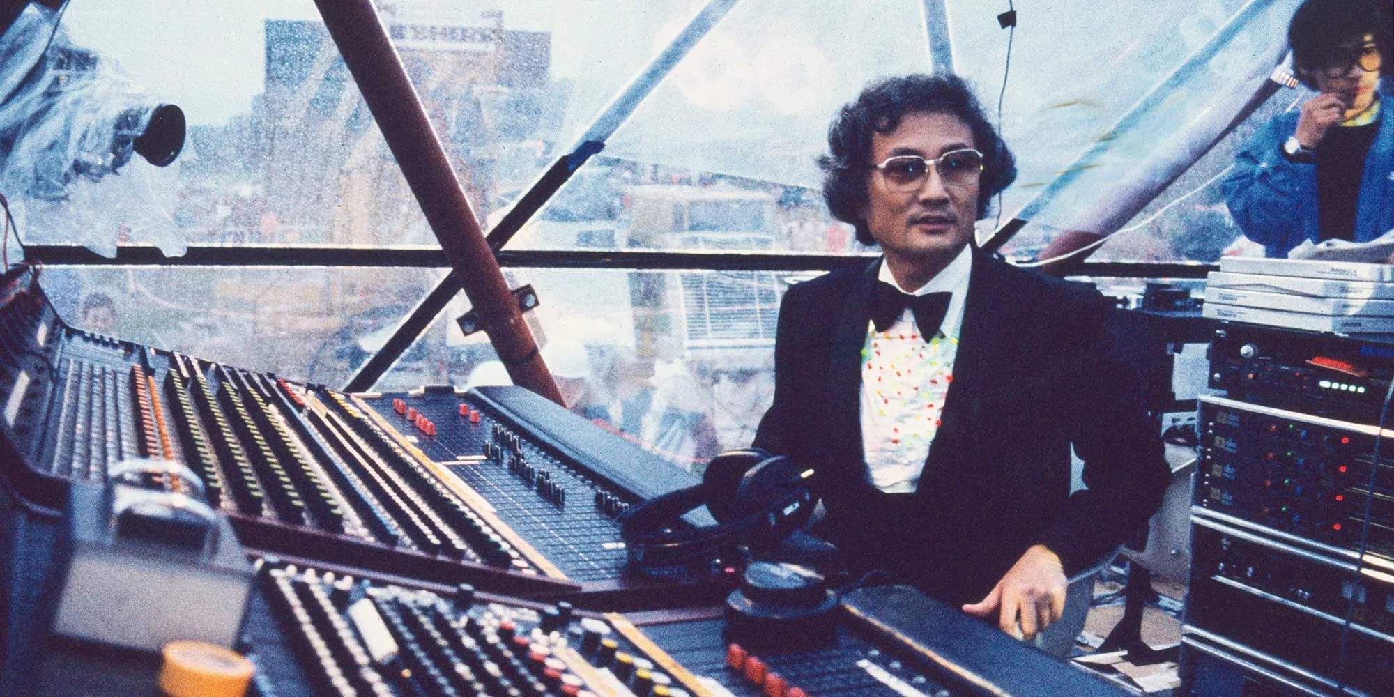 This square photograph shows  Isao Tomita sitting behind a vintage sound mixing board wearing a tuxedo and sunglasses. Sakamoto is fair-skinned with short, dark, wavy hair. He is wearing large, rectangular glasses, a bow tie, and a white shirt with colored confetti dots on it underneath the tuxedo. He looks off to the right with a serious expression. On the sound mixing board are a pair of black headphones and some knobs. Behind the sound mixing board and Sakamoto, a triangular aluminum frame creates a geodesic dome like structure. Behind the frame, people are visible, but they are out of focus. The overall tone of the image is cool with some sepia undertones.