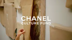 The image shows the text “CHANEL CULTURE FUND” in white lettering superimposed over a scene of artworks being unpacked. There are several artworks covered in brown paper, suggesting that they have been recently transported or stored. One artwork is partially unwrapped, revealing the edges of what appears to be a historical portrait of a person with dark hair and clothing with dark features. A hand is visible, gently peeling back the brown paper to reveal the artwork beneath. The background is a plain, white wall, typical of art galleries or storage spaces. The artwork being unwrapped seems to be encased in a clear plastic or protective layer. The lighting is soft and diffused, creating a sense of care and preservation surrounding the artworks.