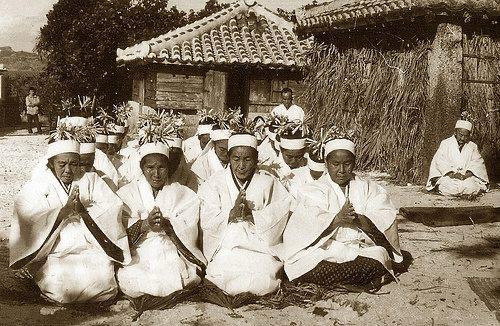A sepia-toned, medium shot depicts a group of people kneeling in neat rows in an outdoor setting. The people, dressed in all-white attire, seem to be taking part in a religious or ceremonial occasion. They have similar headwear with plants in the top.  These individuals are arranged in a semi-circle. Their hands pressed together in a gesture of prayer or respect. The lighting in the shot is soft and diffused. A building is in the background, featuring a tiled roof, and a small structure behind them. Trees are visible beyond the building on the left. The color palette is monochromatic, with subtle variations in tone. This adds to the overall somber and respectful mood of the scene. The ground looks like sand, and some people are in the background standing.