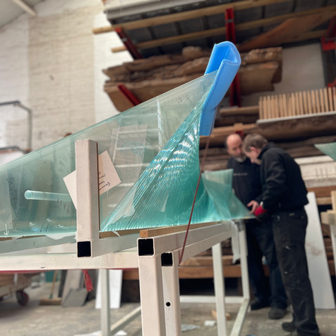 A person in a blue fleece is carefully working on a piece of glass on a carpeted surface. The glass has an irregular shape and is secured in a few places with blue tape. A black glass cutter is in use, held in their hand.  Several tools are visible on the carpet, including a wooden-handled hammer with a metal head, a pair of nipping pliers with brown handles and a dark metal head, and another thin brass tool with a long cylinder body and a brass tip. A small white saucer is visible. The scene is well-lit with overhead lighting, suggesting a workshop or studio environment.