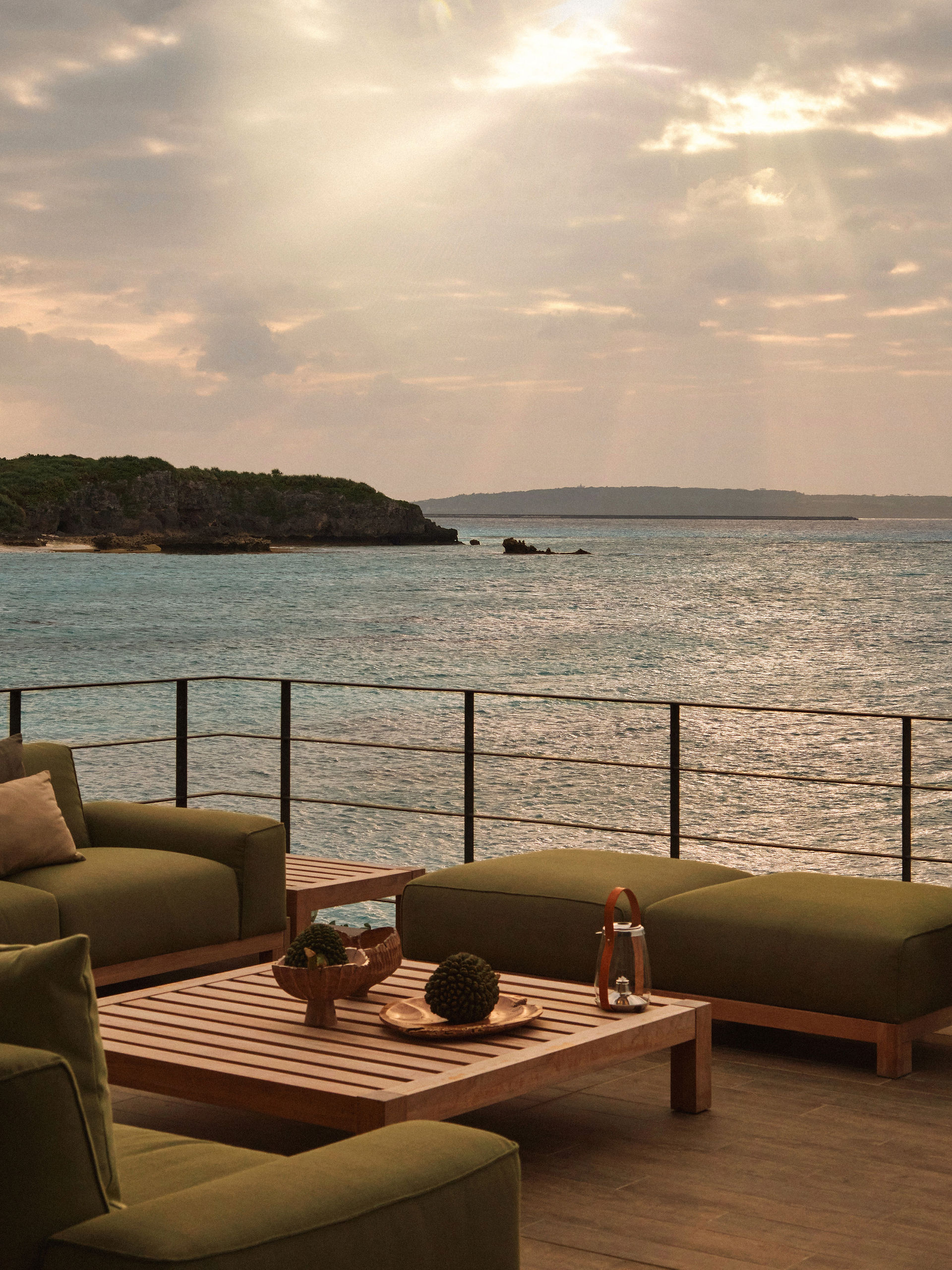 From a high vantage point, a view of a sea scene is offered, with the composition divided into three horizontal sections. The foreground displays a portion of an outdoor seating arrangement. An olive-green armchair is visible on the left, and two ottoman-like cushions of the same color appear on the right, with a wooden table placed between them. A railing that spans the width of the view is situated between the seating and the body of the sea.  Extending into the middle ground is the turquoise sea, its surface exhibiting slight ripples, indicative of a gentle breeze. In the upper-left portion of the middle ground, a small, jagged landmass is visible, covered with green foliage on top and showing sharp, rocky cliffs towards the sea. Directly in the middle of the sea, a smaller cluster of rocks emerges from the water.  The horizon line in the background is composed of a larger, darker landmass stretching across the view, and a sky filled with low-hanging, gray clouds. The overall lighting suggests either dawn or dusk, with a soft, diffused light casting a gentle glow over the scene. The color palette is subdued, with a mix of earth tones, greens, and blues creating a tranquil, serene atmosphere.
