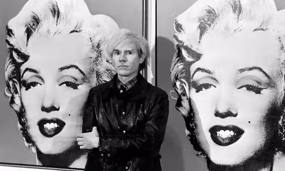 In a black-and-white medium shot, Andy Warhol stands in front of two Marilyn Monroe paintings. Warhol stands in the center, facing directly at the camera, and he has shoulder-length and straight platinum blonde hair that's parted to the side of his head. He has thin lips and a somewhat oblong face. He's wearing a dark color jacket, which is slightly open, and a patterned shirt beneath. His arms are crossed, and his left hand is visible. The two Monroe paintings are Warhol's pop-art style. Monroe's features are painted with broad strokes of black and white, capturing her portrait with an emphasis on her eyes, lips, and the beauty mark near her mouth. Both paintings appear slightly cropped, revealing only partial faces, and are set against neutral tones. The shot is evenly lit, and the focus is sharp, emphasizing both Warhol and his artwork.