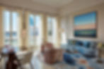 Elegant living room with blue sofa, colorful cushions, and a large ocean painting. Sunlit windows, patterned curtains, and a round table. The Presidential Suite © The St Regis Venice
