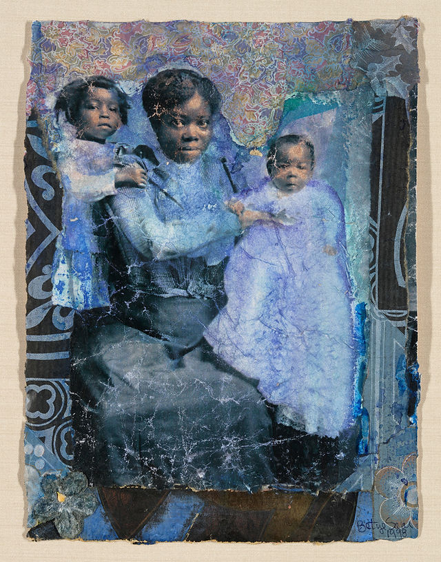 A medium shot of an art piece depicting a woman sitting with two young children, all appearing in shades of blue, black, and white. The art piece is built with heavy layers of what appears to be paper and/or cloth. The woman has a stern expression and appears to sit upright with one child on her lap and another on her left side. The child on her lap is a baby and the child on her left side stands with their right arm around her neck. The appearance of the paper and cloth, as well as the coloring suggests that the art piece is meant to look distressed. The background of the piece features a dark patterned cloth or paper with a light paisley pattern above the woman and children. The right side of the photograph features another dark patterned material. 