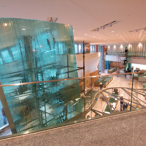 The interior of a large building is shown from an elevated perspective. Construction is in progress, as evidenced by scaffolding, exposed walls, and workers in the space.  To the left of the frame, there's a curved structure constructed from thin, stacked layers of light blue glass. Its arrangement creates a textured and translucent effect.  In the center of the image, there's a scaffolding setup supporting a rounded white structure. Light-colored wood is visible around this area, suggesting ongoing work.  Further into the space, the interior is spacious with a high ceiling illuminated by recessed lighting. In the background to the right, workers can be seen on scaffolding near partially constructed walls. Pallets of materials and tools are scattered around, indicating the ongoing building process.  The viewpoint is from an upper floor behind a glass railing, offering a view into the construction site below. The floor is speckled, adding a textural element to the foreground.