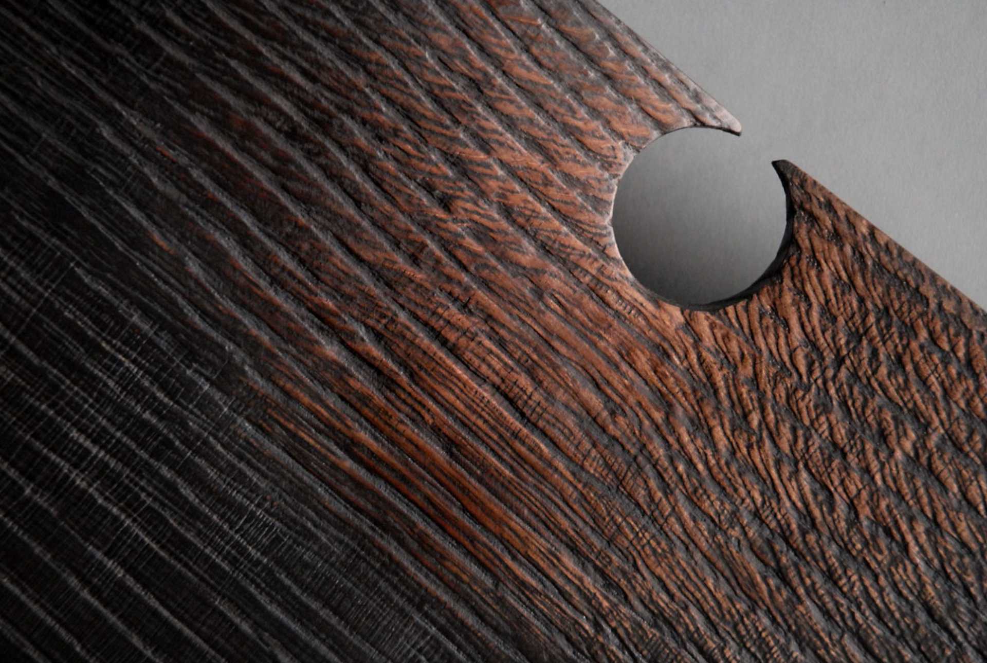 A close-up shot reveals a section of a wooden board, angled to fill the frame diagonally from lower left to upper right. The wood exhibits a rich, dark brown hue, interspersed with black streaks that accentuate its natural grain. This grain pattern is not smooth but textured, featuring pronounced ridges and grooves that run parallel to each other, adding a tactile quality to the surface.  A distinct circular cutout is present on the right edge of the board, revealing a neutral gray background. The edges of this cutout are smooth and precise, contrasting with the rough, grainy texture of the wood. The lighting emphasizes the texture, casting shadows within the grooves and highlighting the raised portions of the wood grain.