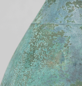 This close-up shot reveals the weathered surface of a metal structure, primarily showcasing a conical or dome-shaped segment. The metal exhibits a striking verdigris patina, a result of copper oxidation, where areas of blue and green hues dominate. These colors are not uniform, instead they form a mosaic-like pattern with variations in shade and texture, suggesting varying degrees of oxidation or exposure to environmental elements.  The surface has a matte finish with a slightly rough, pitted texture, suggesting the material has aged naturally over time and has not been treated with a sealant or coating. The presence of an indiscernible seam or joint in the surface indicates that the structure is pieced together from multiple segments.  There's a stark contrast between the aged metal and the neutral, light gray background, which makes the metal's colors and details more prominent. The lighting is soft and diffused, revealing textures and gradients without harsh shadows. The visual composition focuses on the metal's surface, inviting the viewer to inspect the aging process and the material qualities closely.