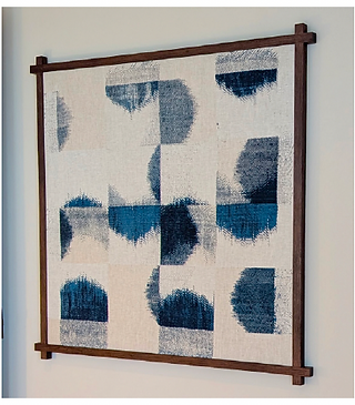 The photograph shows a framed piece of modern art hanging on a pale, neutral wall. The artwork itself features a geometric pattern of overlapping curves and squares in shades of deep blue and off-white, creating a sense of depth and movement. The texture of the fabric adds a tactile quality, with the design appearing slightly blurred or "impressionistic." The fabric is stretched taut within a simple, dark wood frame, which complements the artwork's clean lines and muted color palette. The frame's corners are reinforced with small wooden blocks, adding a subtle architectural detail. The lighting in the photograph is soft and diffused, casting gentle shadows that accentuate the textures and colors of the artwork. The artwork is positioned at a slight angle, which allows the viewer to appreciate its depth and dimensionality.