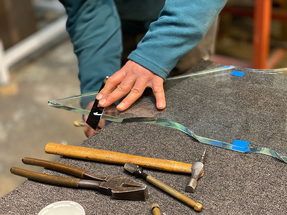 A person in a blue fleece is carefully working on a piece of glass on a carpeted surface. The glass has an irregular shape and is secured in a few places with blue tape. A black glass cutter is in use, held in their hand.

Several tools are visible on the carpet, including a wooden-handled hammer with a metal head, a pair of nipping pliers with brown handles and a dark metal head, and another thin brass tool with a long cylinder body and a brass tip. A small white saucer is visible. The scene is well-lit with overhead lighting, suggesting a workshop or studio environment.