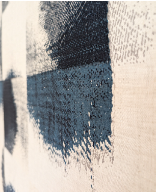 A close-up, slightly blurred, medium shot depicts a section of a textile artwork or fabric design against a solid cream background. The piece features blocks of color applied in a textured, almost faded style, giving the impression of a print on woven material rather than solid painted areas.  In one area, a block of dark blue-gray shades transitions into a muted teal, creating a layered effect of color depth. The application resembles brushstrokes or a diluted dye impact, hinting at an imperfect finish that adds to the piece's handmade appeal.  The texture of the fabric is visible beneath the colored areas, and one can notice faint patterns or the physical weave of the cloth, implying it may be made from natural fibers such as cotton or linen. This texture is particularly apparent where the color is less dense, suggesting that the material underneath influences how the colors appear. The cream background contrasts with the darker shades, providing a neutral field that allows the colors to stand out.