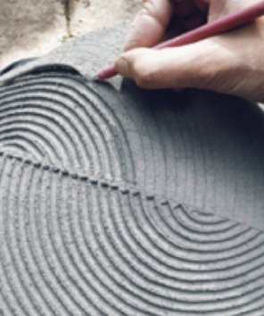 A close-up shot depicts a hand using a stylus to draw concentric semi-circles in a fine-grained surface. The background appears blurred and not in focus. The hand is right-handed, with fair skin, and is holding a slim red stylus. The stylus is creating a shallow curved line in a grey surface, which appears to be a fine-grained material like wet cement or smoothed clay. The lines are neatly arranged in concentric semi-circles, divided by a horizontal dotted line through the center of the piece. The semi-circles span across the piece. The top of the piece is out of focus, as is the background. The image overall has a handcrafted or artistic feel, suggesting that the item being worked on is a custom creation.