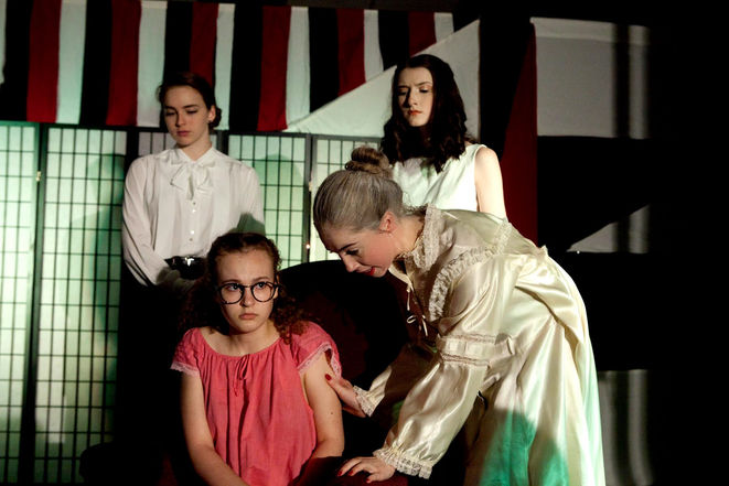 Grace as Rosalie, wearing a pink nightgown and big brown glasses, looks guiltily at an older woman interrogating her as two other women watch.