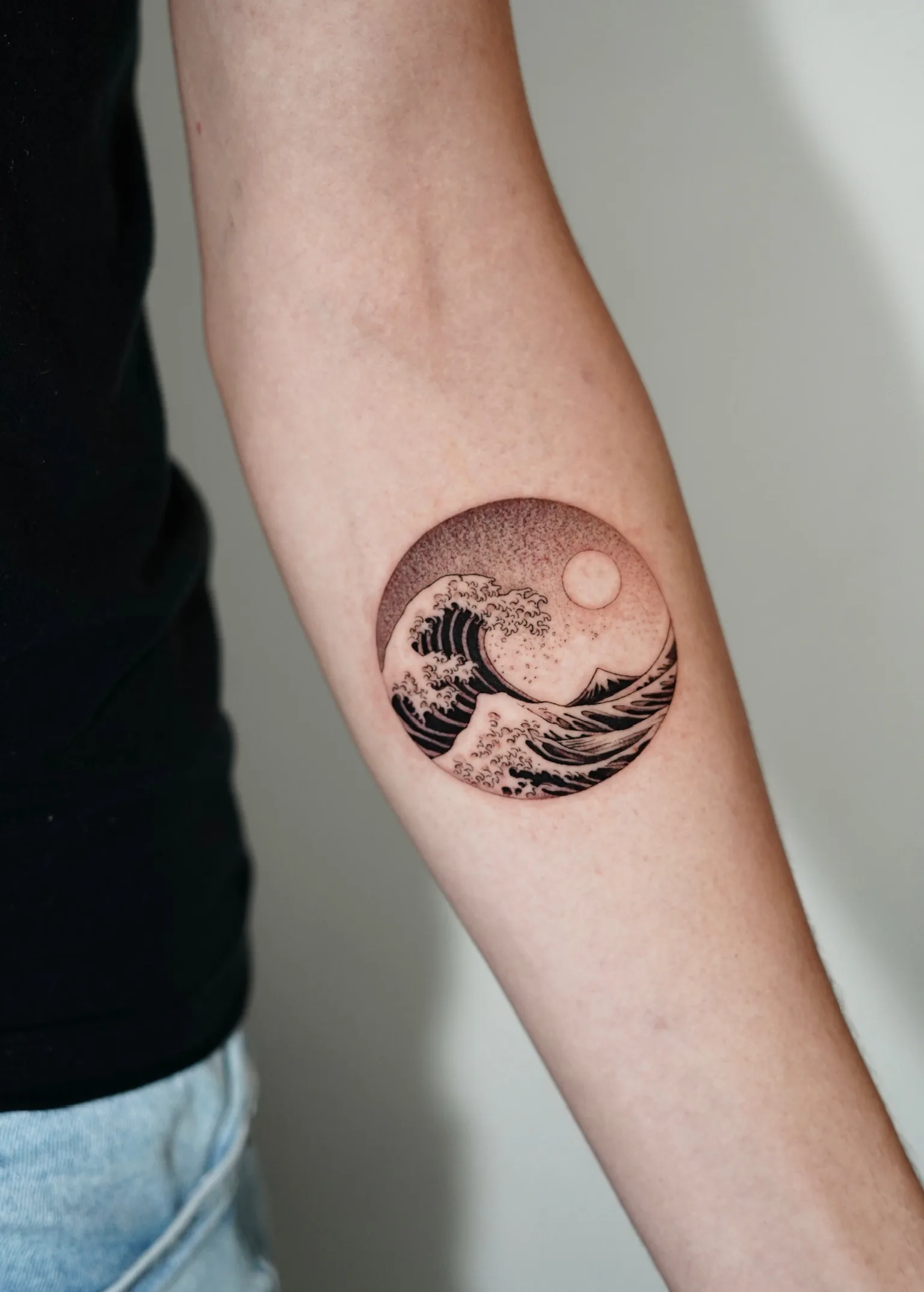 Fineline circular tattoo on the forearm of a Japanese-style great wave with a glowing sun in a dotwork sky, enclosed in a round frame. Seoul Ink Tattoo, Gangnam, Seoul, South Korea.