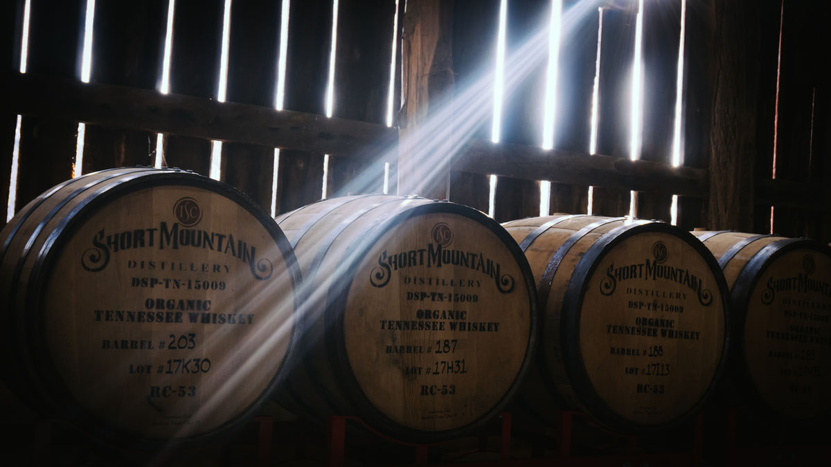 Short Mountain Distillery whiskey barrels inside a dark wooden barrel house with shafts of sunlight