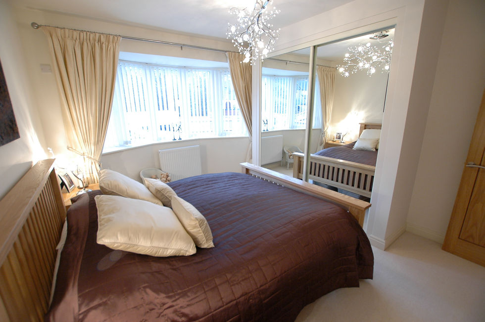 A full image of a cosy, traditional style bedroom. There is a king size oak framed bed and a large bay window. At the foot of the bed is a built-in wardrobe with 2 large mirror doors.