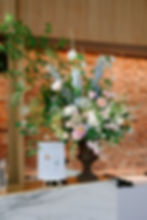 Floral arrangement with pink and white roses in ornate vase, set against a brick wall. Sign reads "Signature Drinks." Bright and elegant.