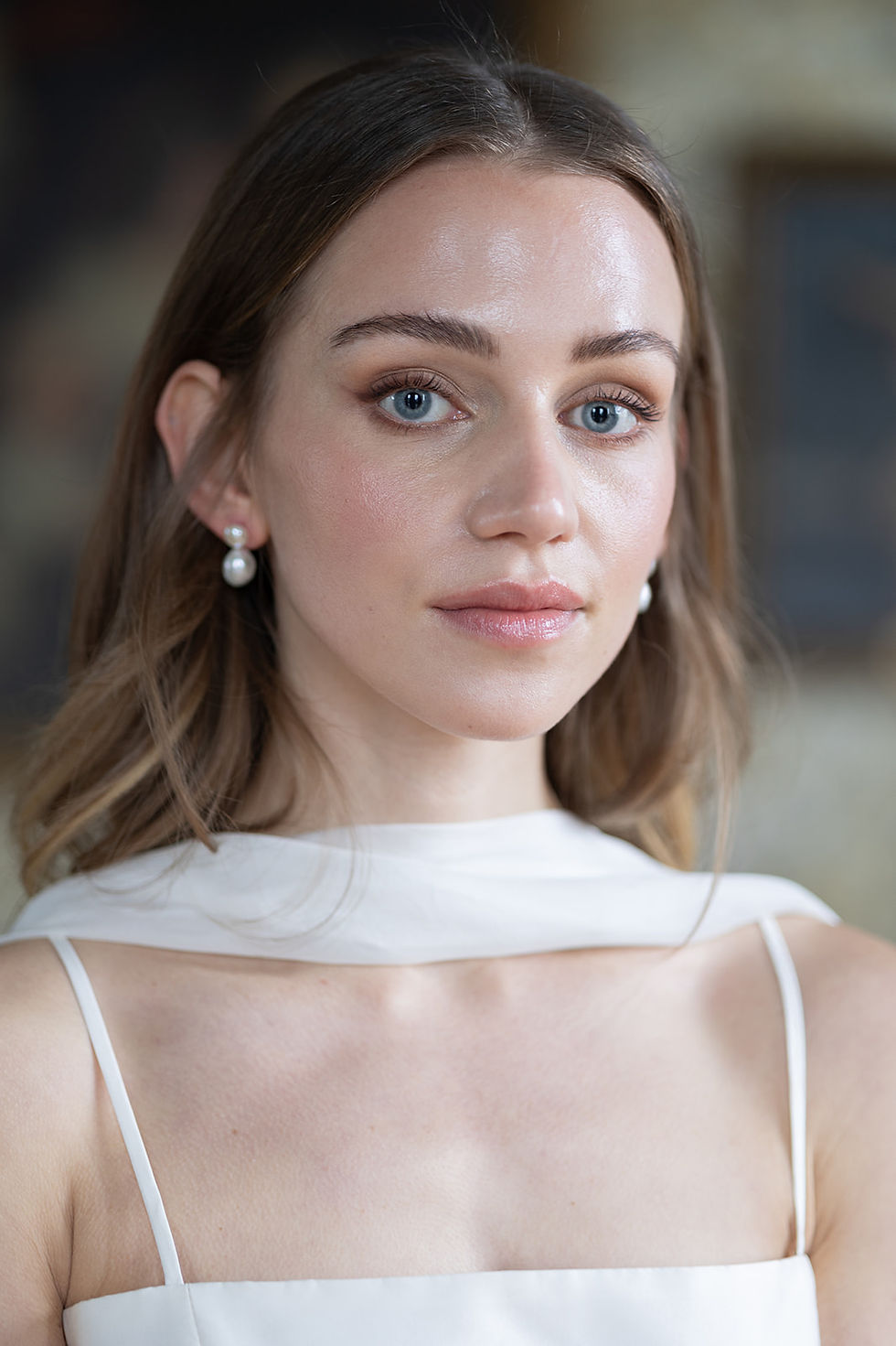 Young woman with long hair and pearl earrings looks at the camera. She's wearing a white dress. Soft, blurred background hints at an interior setting.