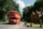 A red double-decker bus labeled "Wedding Special" travels on a road beside a historic brick building, surrounded by lush green trees.